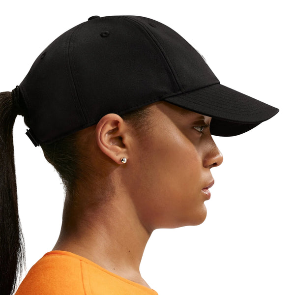 En person set i profil bærer en Nike ACG Club Unstructured Cap i sort/sort, små ørestikker og en orange skjorte. Håret er sat op i en lav hestehale foran en ensfarvet, lys baggrund.