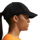 En person set i profil bærer en Nike ACG Club Unstructured Cap i sort/sort, små ørestikker og en orange skjorte. Håret er sat op i en lav hestehale foran en ensfarvet, lys baggrund.