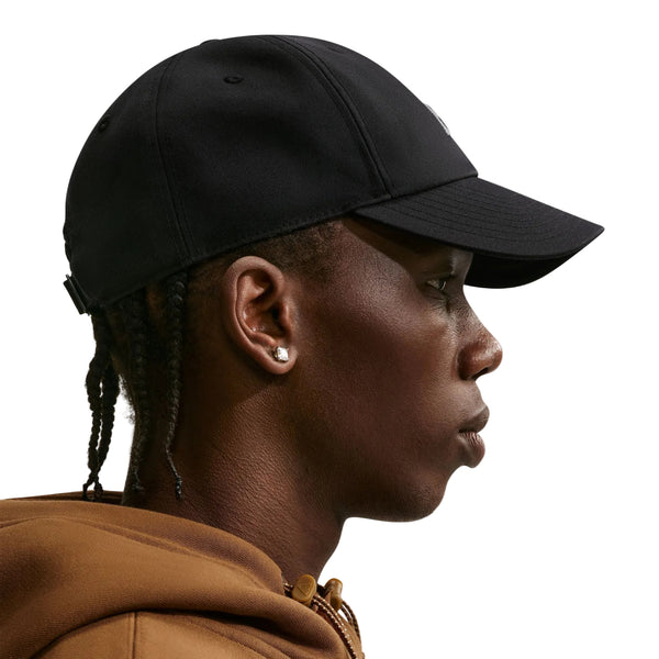 En person set i profil bærer en Nike ACG Club Unstructured Cap i farven sort/sort, en brun hættetrøje og ørestikker foran en ensfarvet hvid baggrund.