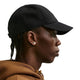En person set i profil bærer en Nike ACG Club Unstructured Cap i farven sort/sort, en brun hættetrøje og ørestikker foran en ensfarvet hvid baggrund.