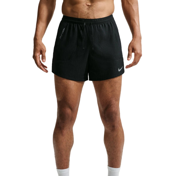 En mand står med ansigtet vendt fremad iført sorte Nike Aero-FIT 5" løbeshorts med indvendig underbuks (herre) i farven sort/reflekterende sølv. Løbeshortsene er udstyret med ventilationsteknologi, snøre i taljen og Nike-logoet på venstre ben. Kun hans overkrop og ben er synlige.