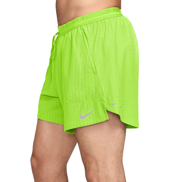 En person har Nike Aero-FIT 5" løbeshorts med indvendig underbuks til mænd i farven Volt/Reflective Silver på. Shortsene har en elastisk linning med snøre, en slids i siden og det karakteristiske Nike-logo. Kun den nedre del af overkroppen og lårene er synlige.
