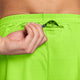 Et nærbillede af en persons hånd, der åbner lynlåsen på den lille baglomme på et par lysegrønne Nike Aero-FIT 5" løbeshorts med underbukser til mænd i farven Volt/Reflective Silver. Den ribstrikkede linning er trykt med teksten »RUNNING«, og shortsene har et struktureret design.