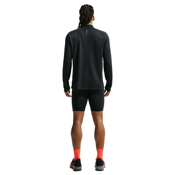 En person står med ryggen til og har en sort Nike Aero-FIT 1/2-Zip løbetrøje, Herre, Sort/Reflekterende Sølv, på, kombineret med sorte, tætsiddende shorts, sorte løbesko med hvide såler og lysorange sokker. Baggrunden er hvid, og personens hår er sat op i lange fletninger. Den åndbare løbetrøje sikrer komfort under bevægelse.