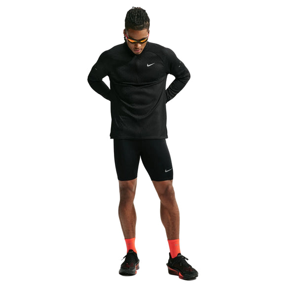 En mand står med blikket let vendt nedad og hænderne bag ryggen. Han er iført en sort Nike Aero-FIT 1/2-Zip løbetrøje til mænd i farvekombinationen sort/reflekterende sølv samt sorte Nike-shorts, orange sokker, sorte sneakers og gule solbriller, alt sammen foran en hvid baggrund.