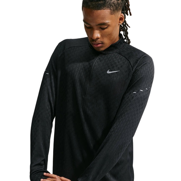 En person med langt, flettet hår har en sort Nike Aero-FIT 1/2-Zip løbetrøje på, Herre, Sort/Reflekterende Sølv – en åndbar løbetrøje med en kvart lynlås. Det reflekterende sølvfarvede Nike-swoosh-logo ses på venstre bryst. Personen kigger ned med armene over kors.