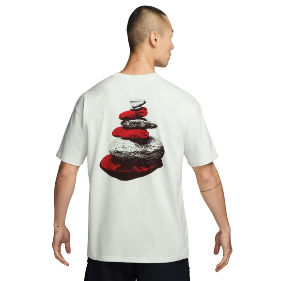 Man ser en person bagfra iført en Nike ACG Dri-FIT T-shirt, Herre, Summit White. På bagsiden af T-shirten er der et grafisk motiv af stablede sten i rødt og gråt, og med armene afslappet ned langs siden fremviser personen denne stilfulde outdoor-T-shirt, der er perfekt til aktive eventyr.