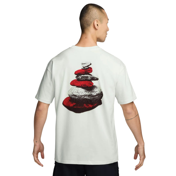 Man ser en person bagfra iført en Nike ACG Dri-FIT T-shirt, Herre, Summit White. På bagsiden af T-shirten er der et grafisk motiv af stablede sten i rødt og gråt, og med armene afslappet ned langs siden fremviser personen denne stilfulde outdoor-T-shirt, der er perfekt til aktive eventyr.