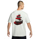 Man ser en person bagfra iført en Nike ACG Dri-FIT T-shirt, Herre, Summit White. På bagsiden af T-shirten er der et grafisk motiv af stablede sten i rødt og gråt, og med armene afslappet ned langs siden fremviser personen denne stilfulde outdoor-T-shirt, der er perfekt til aktive eventyr.