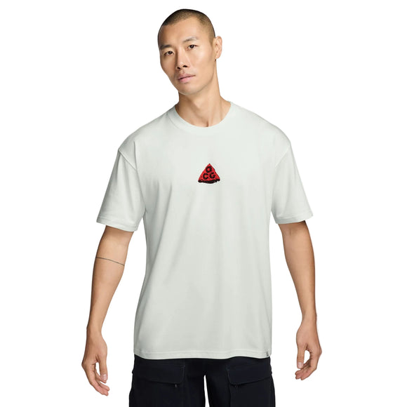 En person med kort hår har en ensfarvet Summit White Nike ACG Dri-FIT T-shirt på, hvor der er et lille rødt trekantlogo placeret midt på brystet. Personen står foran en hvid baggrund med et neutralt udtryk i ansigtet.