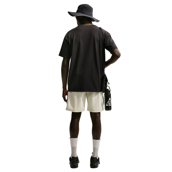 En person står med ryggen til og har en sort Nike ACG Dri-FIT T-shirt, Herre, på, kombineret med lyse shorts, hvide sokker og sorte sko. Personen holder en sort vandflaske og har en sort taske hængende over den ene skulder, alt sammen foran en hvid baggrund.