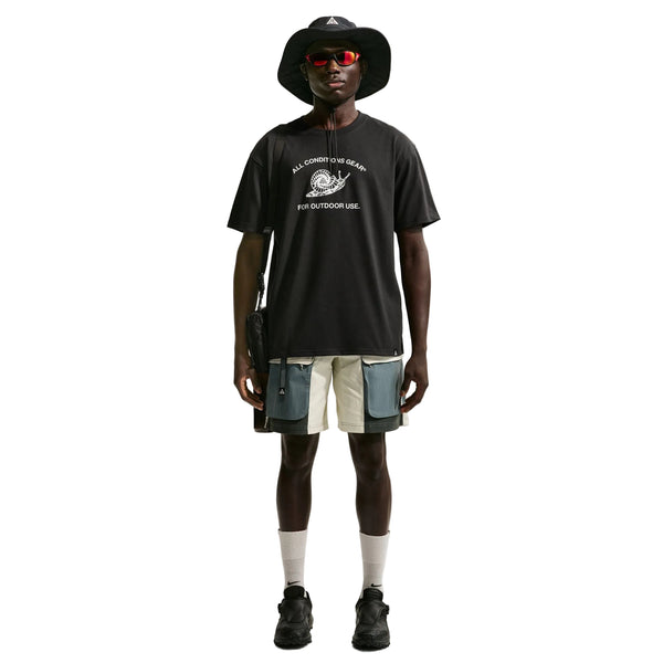En person står med ansigtet vendt fremad foran en hvid baggrund og er iført en sort bucket hat, en Nike ACG Dri-FIT T-shirt (herre, sort), flerfarvede utility-shorts, sorte sneakers, hvide sokker, røde solbriller og en sort skuldertaske.