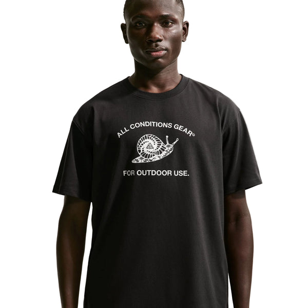 En person har en Nike ACG Dri-FIT T-shirt, herre, sort, på, som er prydet med et hvidt sneglemotiv og teksten »ALL CONDITIONS GEAR FOR OUTDOOR USE« ovenfor og nedenfor. Med den ensfarvede hvide baggrund fremstår denne T-shirt med et rent og moderne look.
