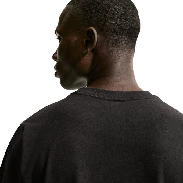 Der ses en person set bagfra og lidt fra siden, iført en sort Nike ACG Dri-FIT T-shirt til mænd. Baggrunden er hvid, og personens ansigt er delvist synligt i profil, mens vedkommende kigger ud mod venstre.