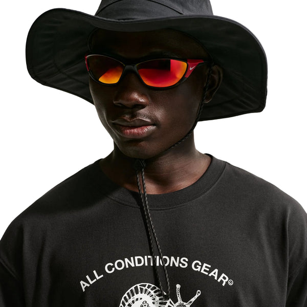 En person iført en sort hat med bred skygge, røde solbriller med spejlglas og en Nike ACG Dri-FIT T-shirt, Herre, Sort, står foran en hvid baggrund.