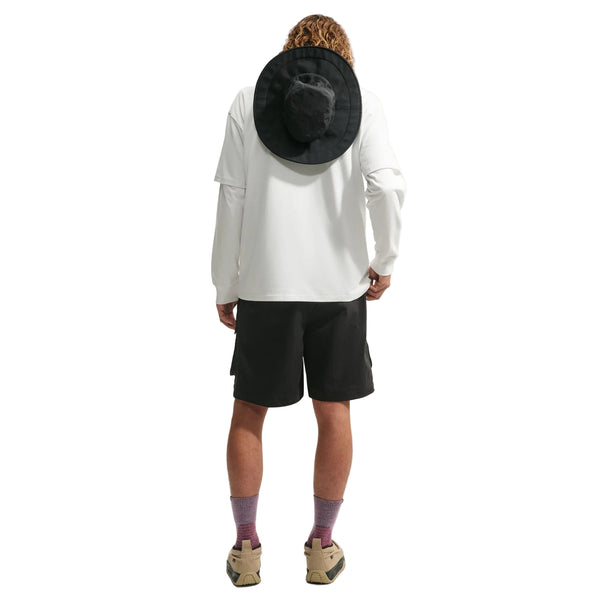 En person med krøllet hår står med ryggen til foran en ensfarvet hvid baggrund og er iført en hvid Nike ACG Dri-FIT T-shirt (Herre, hvid/Summit White), sorte shorts, lilla sokker, beige sko og en sort hat med bred skygge på ryggen.