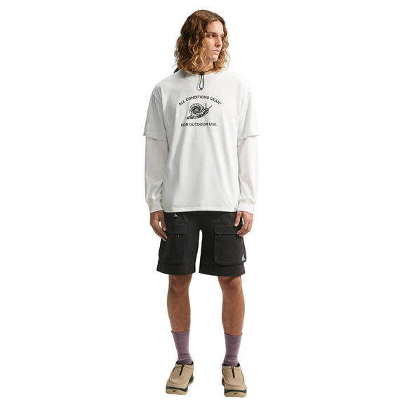 En person med langt, bølget hår har en hvid langærmet skjorte på under en Nike ACG Dri-FIT T-shirt til mænd i farven White/Summit White, kombineret med sorte cargoshorts, lilla sokker og beige sko. Personen står med ansigtet vendt fremad foran en hvid baggrund.