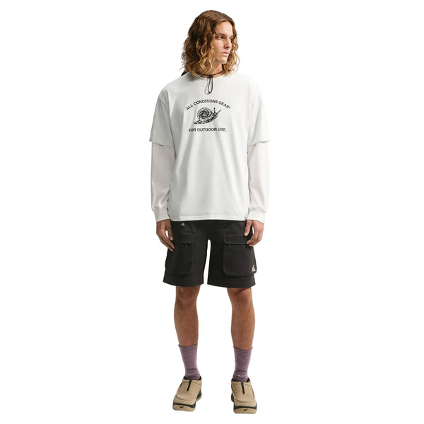 En person med langt, bølget hår har en hvid langærmet skjorte på under en Nike ACG Dri-FIT T-shirt til mænd i farven White/Summit White, kombineret med sorte cargoshorts, lilla sokker og beige sko. Personen står med ansigtet vendt fremad foran en hvid baggrund.