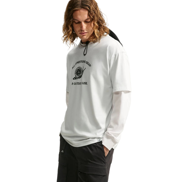 En person med langt, bølget hår er klædt i afslappet herretræningstøj, med sorte bukser og en hvid langærmet skjorte under en Nike ACG Dri-FIT T-shirt til mænd i farven White/Summit White. Den kortærmede outdoor-T-shirt har et motiv af en snegl med tekst. Personen kigger ned med den ene hånd i lommen foran en hvid baggrund.