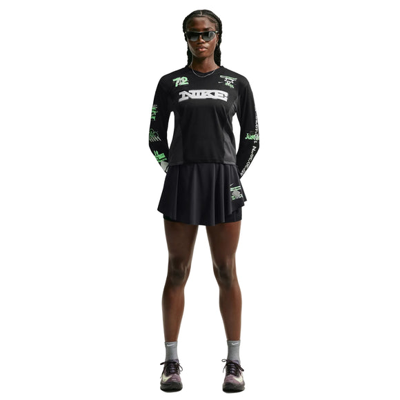 En person står med ansigtet vendt fremad og er iført en sort langærmet T-shirt med grafisk print, en Nike Swift Dri-FIT løbeskirt (sort/sort/reflekterende sølv), grå sokker, sorte sneakers og solbriller. Hænderne er bag ryggen, og håret er flettet.