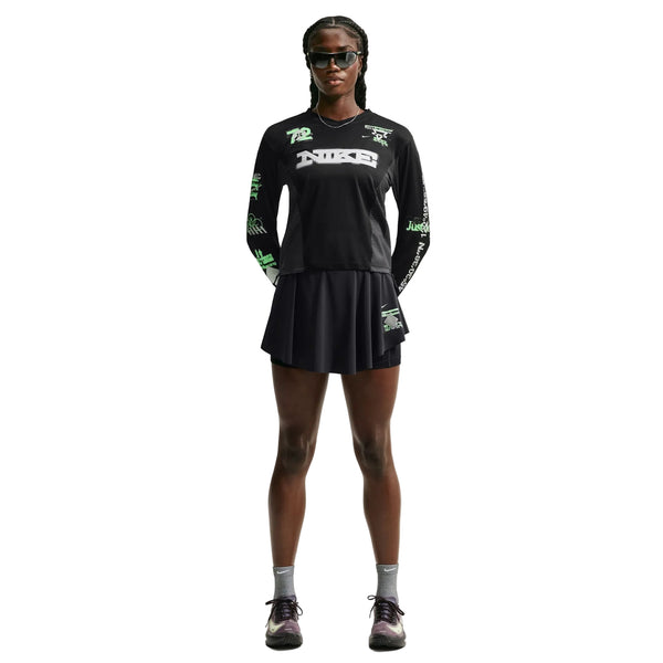 En person står med ansigtet vendt fremad og er iført en sort langærmet T-shirt med grafisk print, en Nike Swift Dri-FIT løbeskirt (sort/sort/reflekterende sølv), grå sokker, sorte sneakers og solbriller. Hænderne er bag ryggen, og håret er flettet.