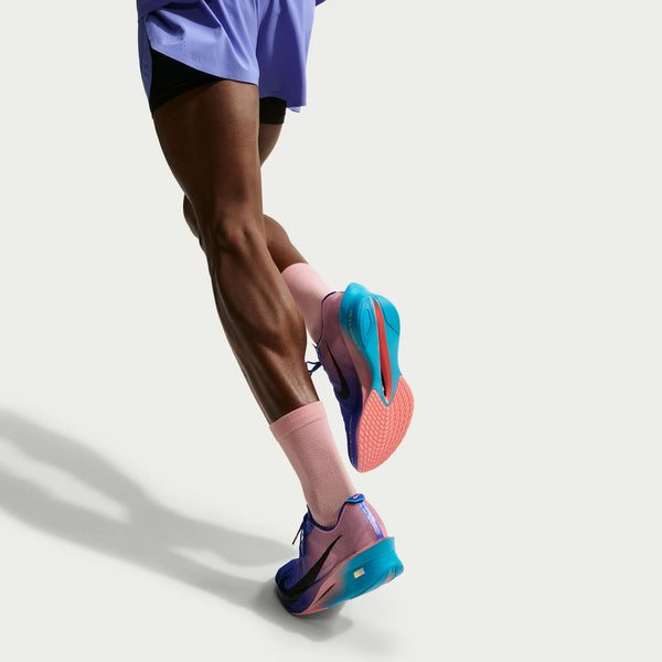 En persons ben fra midten af låret og ned, iført lilla løbeshorts, lyserøde sokker og Nike Vaporfly Next% 4 Herre Racer Blue/White-Blue Lightning-sko, foran en ensfarvet hvid baggrund.