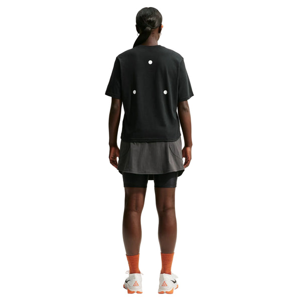 En person står med ryggen til og har en sort Nike ACG Dri-FIT T-shirt Dame på, sammen med sorte shorts, en grå undertrøje, orange sokker og hvide sneakers med orange såler. Baggrunden er helt hvid.