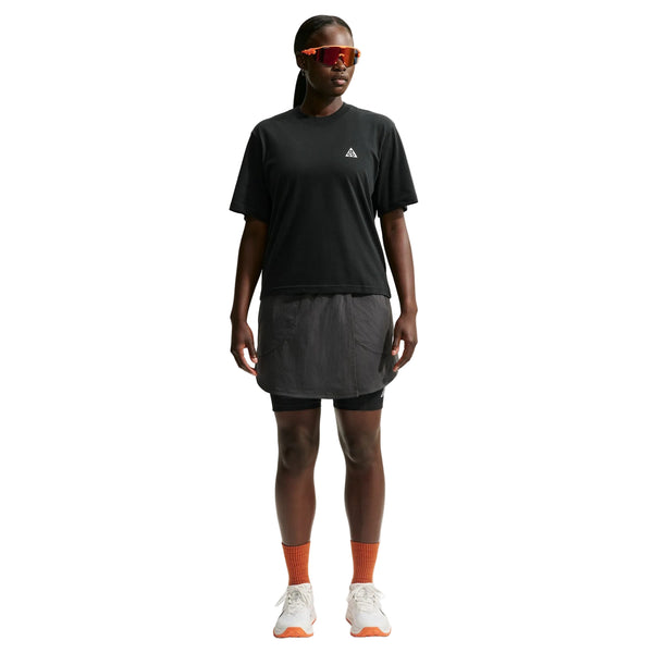 En person står med ansigtet vendt fremad og er iført en sort Nike ACG Dri-FIT T-shirt (Dame), sorte shorts med en overliggende nederdel, orange sokker, hvide sneakers og rødtonede sportsolbriller. Baggrunden er ensfarvet hvid.
