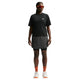 En person står med ansigtet vendt fremad og er iført en sort Nike ACG Dri-FIT T-shirt (Dame), sorte shorts med en overliggende nederdel, orange sokker, hvide sneakers og rødtonede sportsolbriller. Baggrunden er ensfarvet hvid.