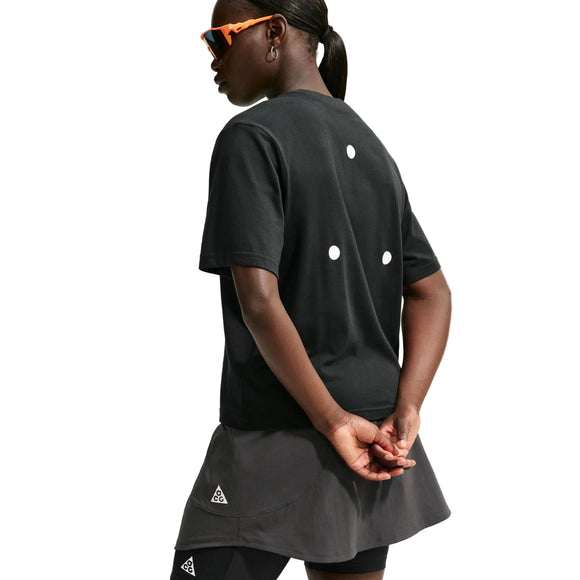 En person iført sorte solbriller, en sort Nike ACG Dri-FIT-T-shirt til kvinder med tre hvide prikker på ryggen samt en sort nederdel med leggings står med hænderne foldet bag ryggen foran en hvid baggrund.