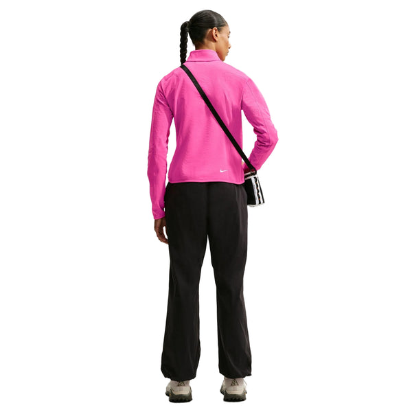 En kvinde står med ryggen til og har en Nike Therma-FIT 1/2-Zip Top til kvinder i farverne Desert Pink/Lt Orewood Brn/Summit White på, kombineret med sorte bukser og lyse sko. Hun bærer en sort skuldertaske, og hendes mørke hår er sat op i en fletning. Baggrunden er ensfarvet hvid.
