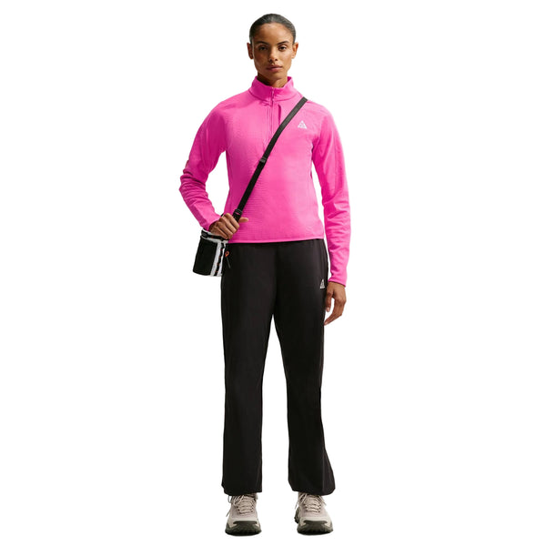 En person står med ansigtet vendt fremad iført en lyserød Nike Therma-FIT 1/2-Zip Top til kvinder i farven Desert Pink, kombineret med sorte bukser, lysegrå sko og en sort skuldertaske. Baggrunden er ensfarvet hvid.