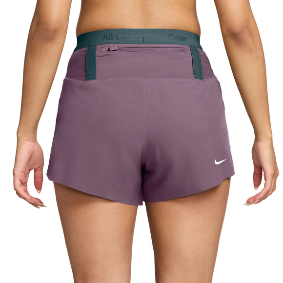 Der vises en person set bagfra, der har Nike ACG Dri-FIT Mid-Rise Trail-løbeshorts med indvendig underbuks til kvinder i farverne Violet Dust/Mineral Slate/Summit White på. Shortsene har en grøn elastisk linning med teksten »All Conditions Gear«, og Nike-logoet er placeret nederst til højre. Hendes arme hænger afslappet ned langs siden, hvilket understreger den behagelige pasform på disse trail-løbeshorts.