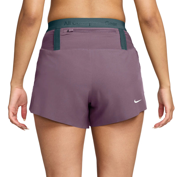Der vises en person set bagfra, der har Nike ACG Dri-FIT Mid-Rise Trail-løbeshorts med indvendig underbuks til kvinder i farverne Violet Dust/Mineral Slate/Summit White på. Shortsene har en grøn elastisk linning med teksten »All Conditions Gear«, og Nike-logoet er placeret nederst til højre. Hendes arme hænger afslappet ned langs siden, hvilket understreger den behagelige pasform på disse trail-løbeshorts.