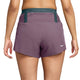Der vises en person set bagfra, der har Nike ACG Dri-FIT Mid-Rise Trail-løbeshorts med indvendig underbuks til kvinder i farverne Violet Dust/Mineral Slate/Summit White på. Shortsene har en grøn elastisk linning med teksten »All Conditions Gear«, og Nike-logoet er placeret nederst til højre. Hendes arme hænger afslappet ned langs siden, hvilket understreger den behagelige pasform på disse trail-løbeshorts.