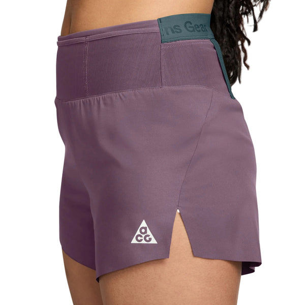 Et nærbillede set fra siden af en person, der bærer Nikes ACG Dri-FIT Mid-Rise Trail-løbeshorts med indvendig underbuks til kvinder i farvekombinationen Violet Dust/Mineral Slate/Summit White. Shortsene har et lille hvidt ACG-trekantlogo på venstre lår og en kort sideslids tæt på sømmen, mens linningen er fremhævet med mørkegrøn (Mineral Slate), hvilket understreger disse stilfulde og højtydende trail-løbeshorts, der er designet med fokus på komfort.