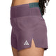 Et nærbillede set fra siden af en person, der bærer Nikes ACG Dri-FIT Mid-Rise Trail-løbeshorts med indvendig underbuks til kvinder i farvekombinationen Violet Dust/Mineral Slate/Summit White. Shortsene har et lille hvidt ACG-trekantlogo på venstre lår og en kort sideslids tæt på sømmen, mens linningen er fremhævet med mørkegrøn (Mineral Slate), hvilket understreger disse stilfulde og højtydende trail-løbeshorts, der er designet med fokus på komfort.