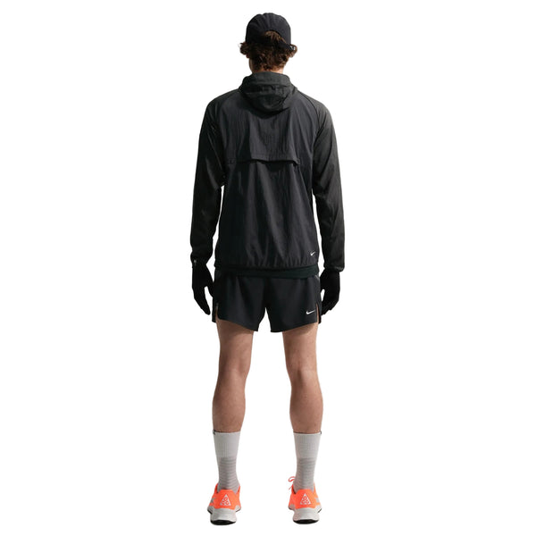 En person står med front mod kameraet iført en sort jakke med hætte, Nike ACG "Second Sunrise" 5" kortforede shorts til mænd i Black/Anthracite/Summit White, en sort kasket, sorte handsker, hvide strømper og lyse orange sportssko på en hvid baggrund.
