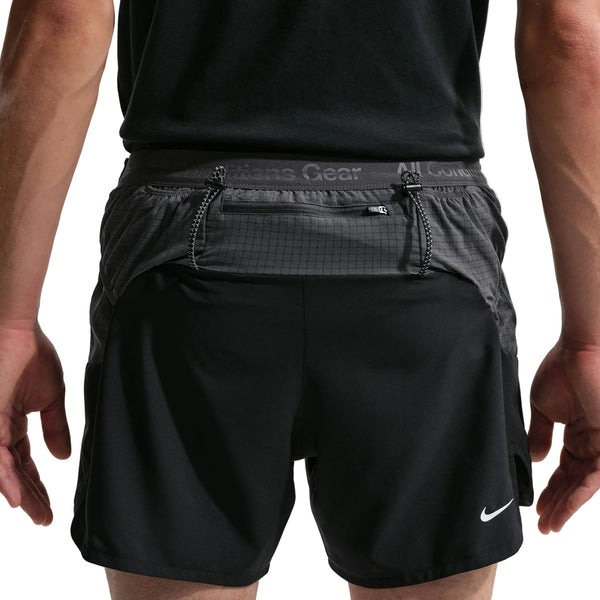 En person, der ses bagfra, er iført sorte Nike ACG "Second Sunrise" 5" Brief-Lined Shorts (Herre, Black/Anthracite/Summit White) med en sort skjorte; shortsene har en lynlåslomme, Dri-FIT ADV-teknologi og synlige syninger.
