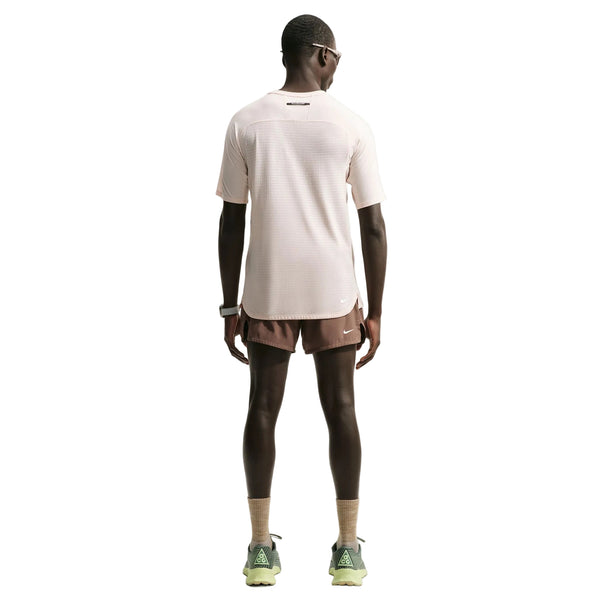 En person står med ryggen til kameraet iført en lyserød Nike ACG Dri-FIT ADV Trail Running Top til mænd i farven Lt Orewood Brn/Summit White, brune sportsshorts, lysebrune sokker, grønne sneakers, et smartwatch og solbriller. Baggrunden er helt hvid.