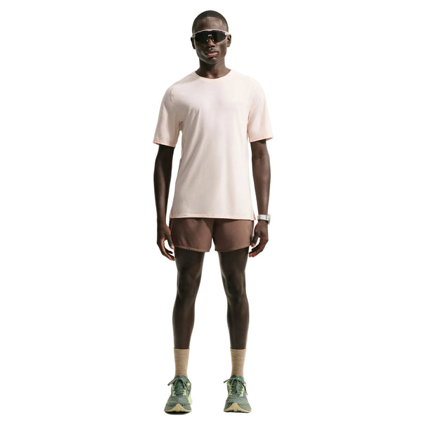 En mand står med ansigtet vendt fremad iført en lysbeige Nike ACG Dri-FIT ADV Trail Running Top, Herre, Lt Orewood Brn/Summit White – ofte kaldet en trailløb-T-shirt til mænd – kombineret med brune shorts, beige sokker, grønne sneakers, sorte solbriller og et armbåndsur, foran en hvid baggrund.