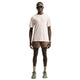 En mand står med ansigtet vendt fremad iført en lysbeige Nike ACG Dri-FIT ADV Trail Running Top, Herre, Lt Orewood Brn/Summit White – ofte kaldet en trailløb-T-shirt til mænd – kombineret med brune shorts, beige sokker, grønne sneakers, sorte solbriller og et armbåndsur, foran en hvid baggrund.