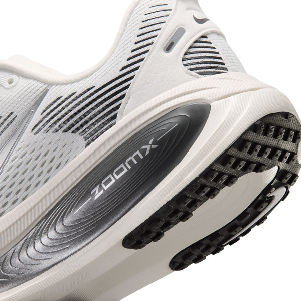 Nærbillede af hælen og sålen på Nike Vomero 18 Dame-løbeskoen i farvekombinationen Summit White/Metallic Silver-Sail, der viser en hvid overdel med sort mønster og ordet ZOOMX på den grå mellemsål. Denne sko har maksimal dæmpning, en overdel i mesh samt detaljer i sort og gråt.