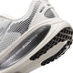 Nærbillede af hælen og sålen på Nike Vomero 18 Dame-løbeskoen i farvekombinationen Summit White/Metallic Silver-Sail, der viser en hvid overdel med sort mønster og ordet ZOOMX på den grå mellemsål. Denne sko har maksimal dæmpning, en overdel i mesh samt detaljer i sort og gråt.