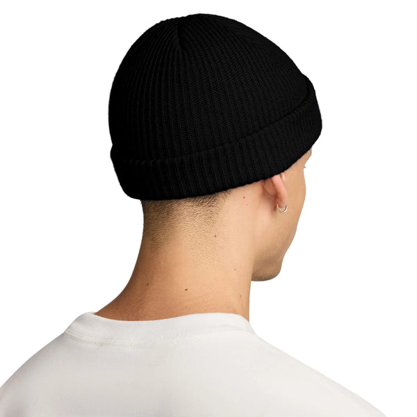 Der ses en person set bagfra, der bærer en sort Nike ACG Terra Beanie – et unisex-tilbehør, der nemt kan kombineres med en frisk, hvid skjorte. Personens korte hår og den lille ringørering i venstre øre tilføjer et diskret stilfuldt touch, mens den ensfarvede hvide baggrund holder fokus på dette ubesværet cool look.