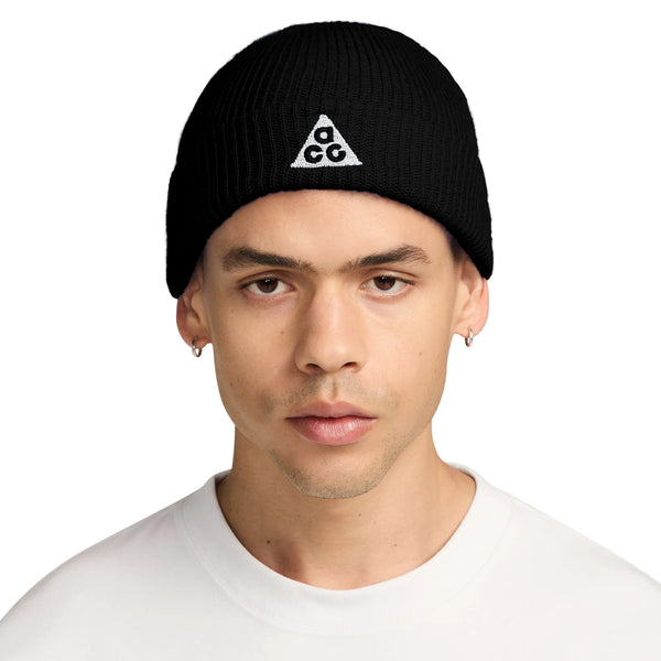 En person iført den sorte Nike ACG Terra Beanie (unisex) med et hvidt trekantet logo samt en hvid T-shirt med rund hals står med ansigtet vendt fremad foran en ensfarvet hvid baggrund. Nike-beanien passer godt til personens korte hår og små ringøreringe.