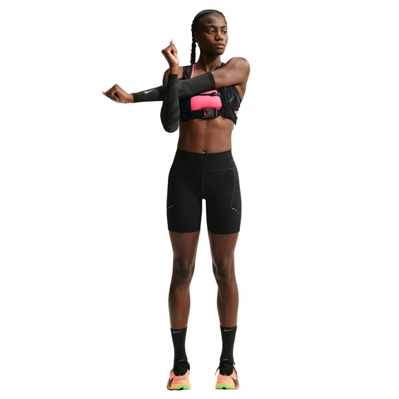 En kvinde i sportstøj strækker sin venstre arm ud over brystet. Hun har en Nike-sport-BH på, højtaljede 7" løbeshorts med lommer til kvinder i sort/reflekterende sølv, sorte sokker og løbesko. Baggrunden er hvid, og hun kigger mod venstre.