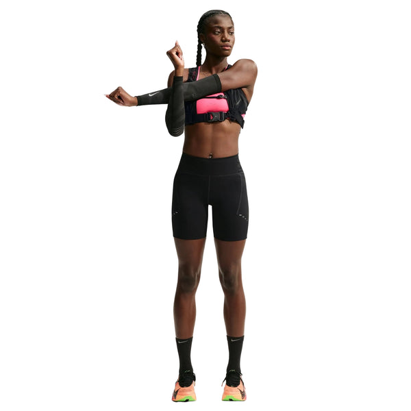 En kvinde i sportstøj strækker sin venstre arm ud over brystet. Hun har en Nike-sport-BH på, højtaljede 7" løbeshorts med lommer til kvinder i sort/reflekterende sølv, sorte sokker og løbesko. Baggrunden er hvid, og hun kigger mod venstre.