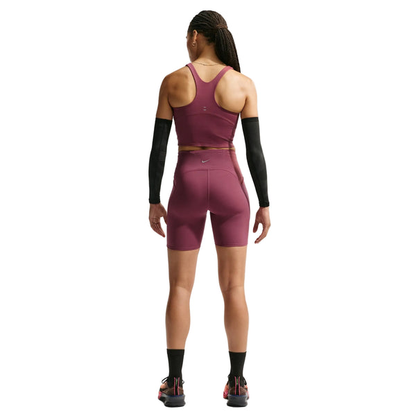 En person står med ryggen til, iført en rødbrun Nike Dri-FIT-sportstrøje og Nike High-Waisted 7″ løbeshorts med lommer til kvinder i farven Pink Smoke/Reflective Silver, kombineret med sorte armvarmere, sorte sokker og sportssko. Baggrunden er ensfarvet hvid.