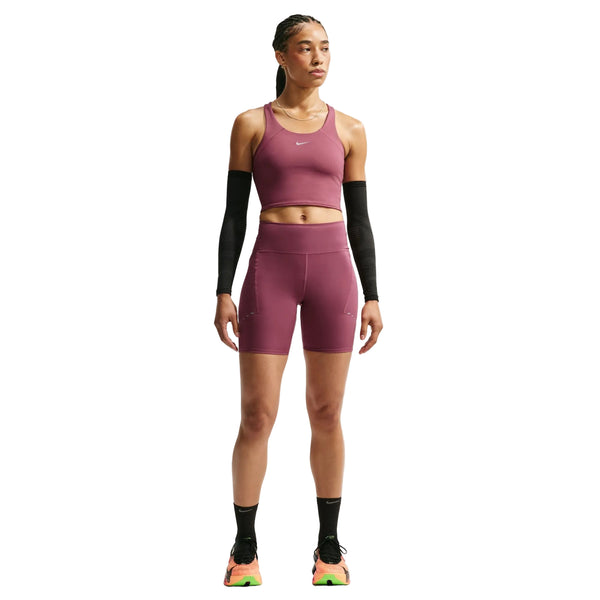 En person står med ansigtet vendt fremad foran en hvid baggrund og har Nikes Dame High-Waisted 7″ løbeshorts med lommer i farven Pink Smoke/Reflective Silver på, en matchende lilla sports-BH, sorte armvarmere, sorte sokker og flerfarvede sportssko.